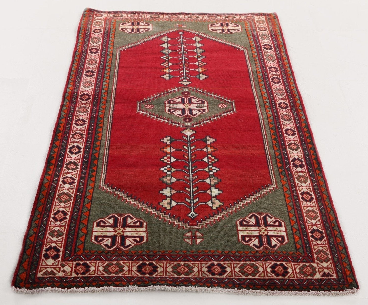 A traditional rectangular wool rug with a red central field, olive green borders, and cream and dark accents in geometric and floral motifs.