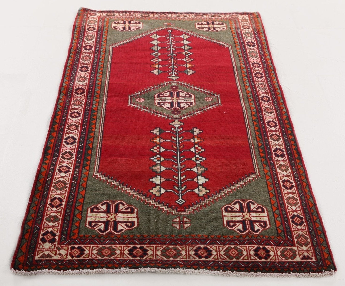 A traditional rectangular wool rug with a red central field, olive green borders, and cream and dark accents in geometric and floral motifs.